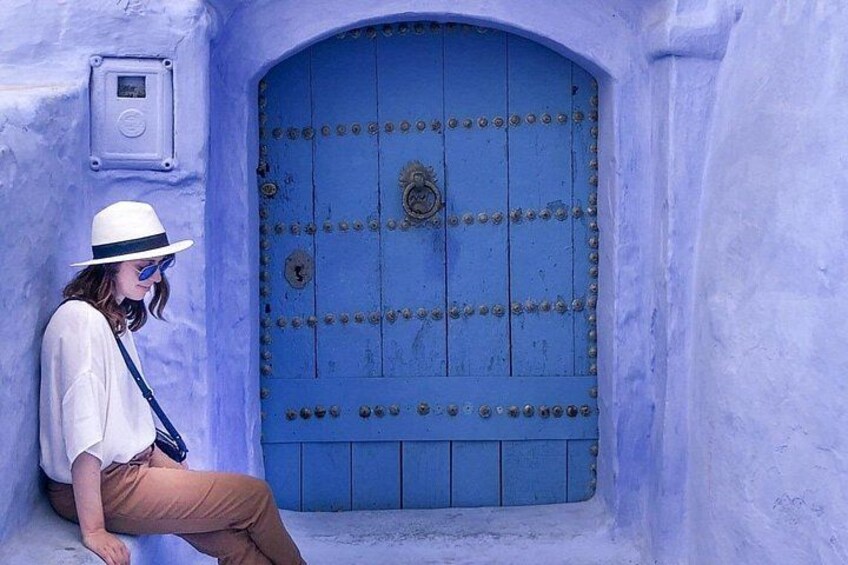A person sitting in front of a blue door with metal studs, wearing a white hat and brown pants. 1