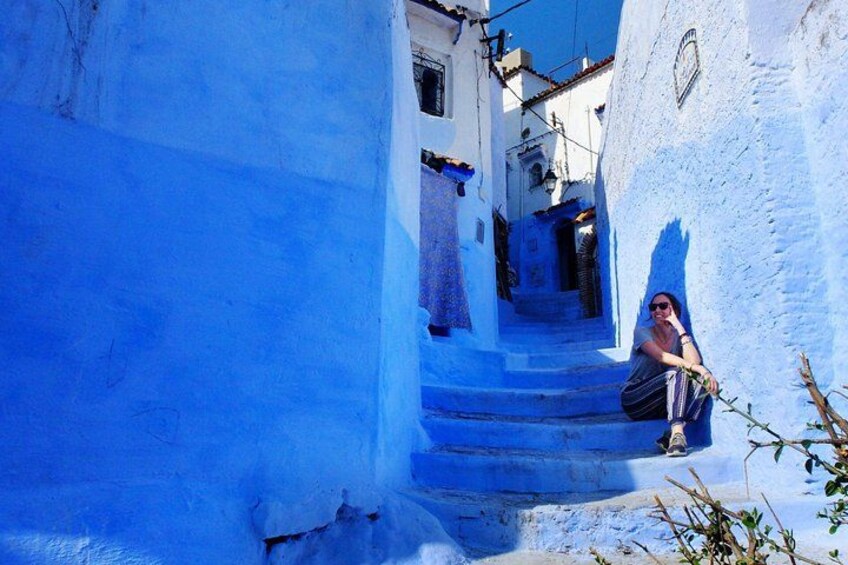 A narrow street with blue-painted buildings, a person sitting on a step, and a staircase leading to a door. 5