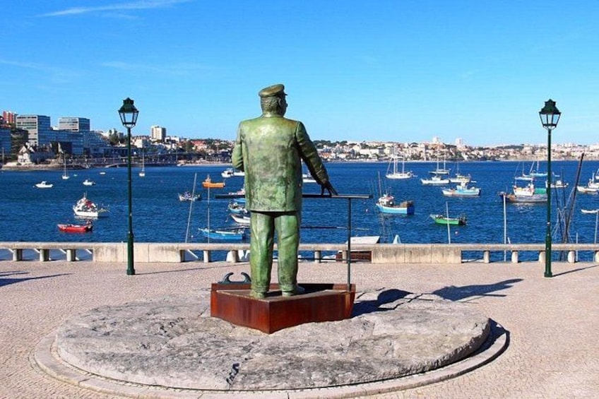 Una estatua de un hombre con traje verde, un puerto con barcos y un paisaje urbano al fondo. 3