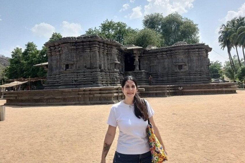 Une personne se tient devant une ancienne structure en pierre, ornée de sculptures complexes, et est entourée d’une zone sablonneuse, avec des arbres en arrière-plan. 1