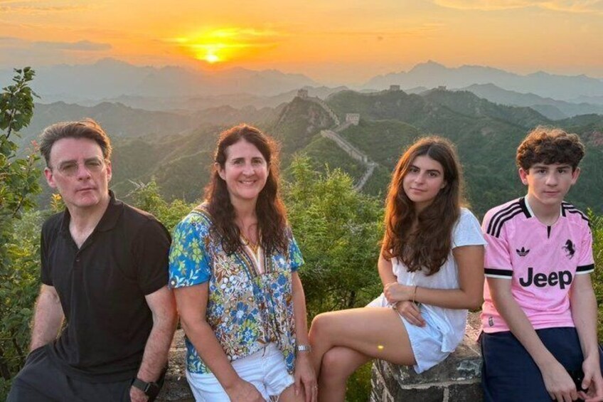 A group of people sitting on a stone ledge with a mountainous landscape and a sunset in the background. 1