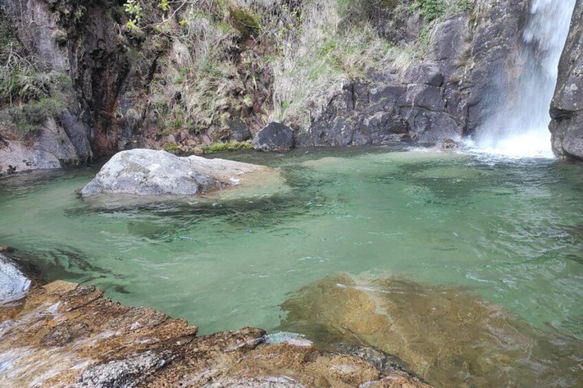 A clear pool with a waterfall, surrounded by rocky terrain and sparse vegetation. 1