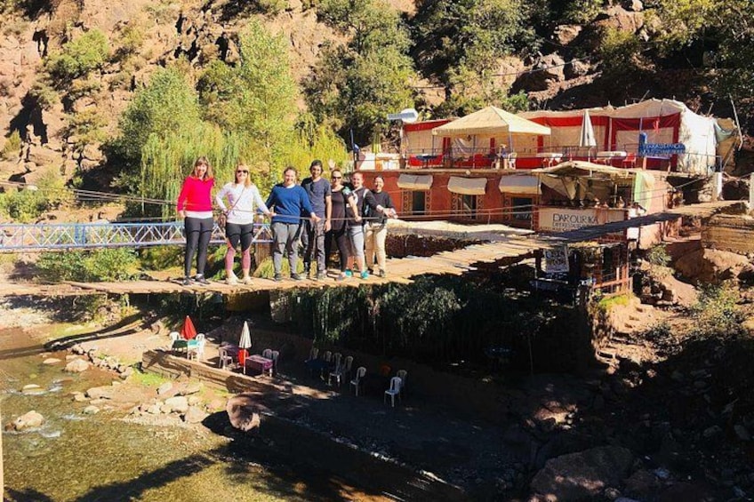 A group of people standing on a wooden bridge over a river, with buildings and greenery in the background. 2