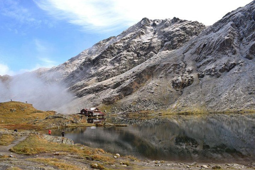 A mountain landscape with a lake, a cabin, and a person standing on a hill. 1