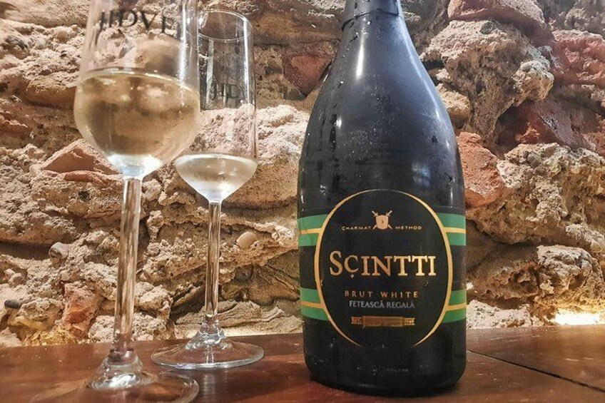 A bottle of Sci.ntti Brut White wine and two glasses on a wooden surface with a rocky background. 3