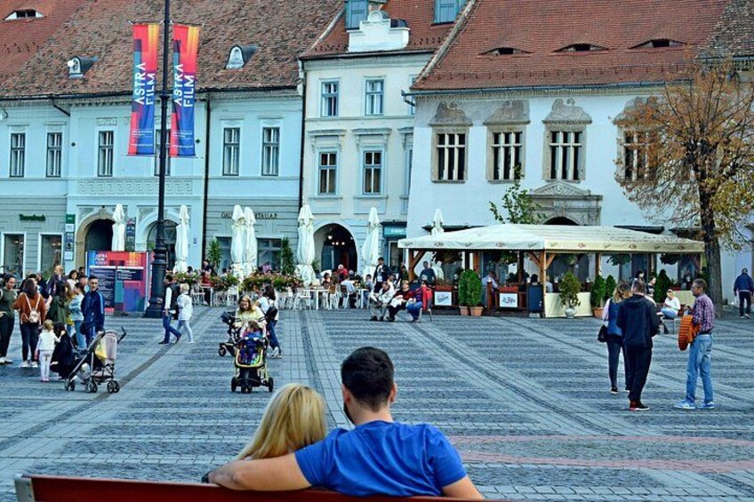Ein belebter Platz mit Fußgängern, ein Café mit Außenbestuhlung und ein Gebäude im klassischen Stil. 1