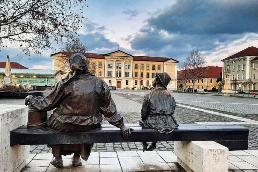 A statue of two figures sitting on a bench, with buildings in the background. 5