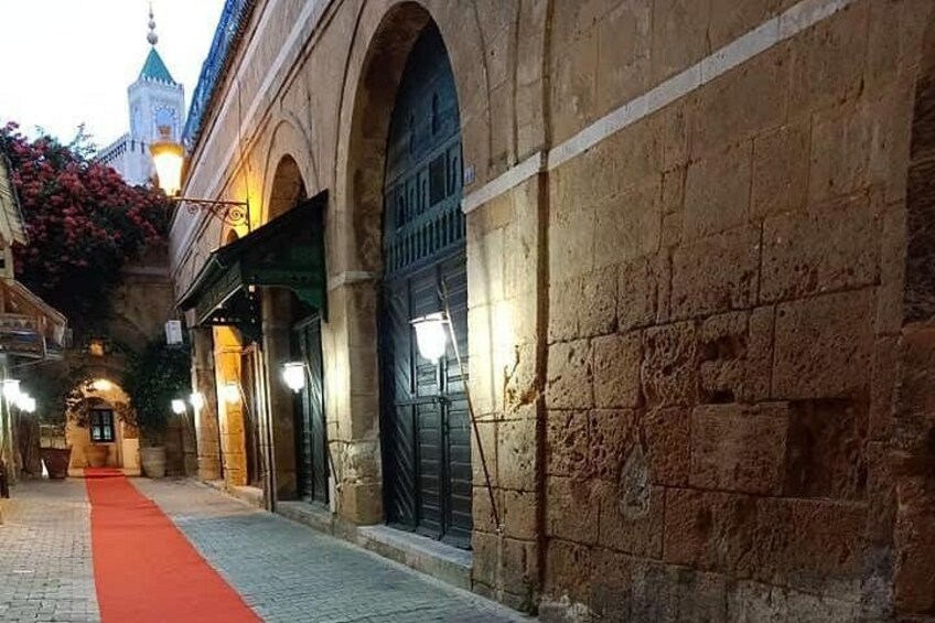 A cobblestone street with a red carpet, stone buildings with arched doorways, and a tower in the background. 3