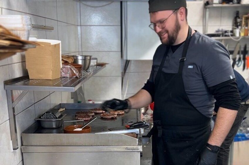 A chef cooking on a grill in a kitchen with stainless steel appliances and shelves with food items. 2