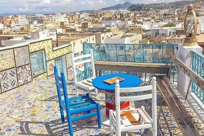 A rooftop terrace with a blue table and chairs, patterned tiles, and a cityscape in the background. 1
