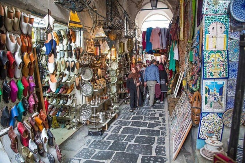 A narrow market street with various stalls selling shoes, pottery, and textiles. 5