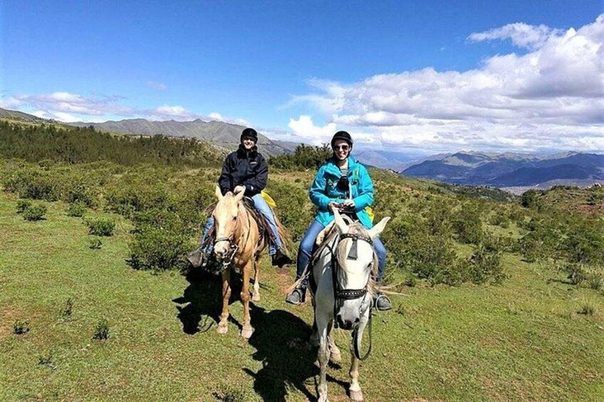 Two people riding horses in a grassy field with mountains in the background. 1