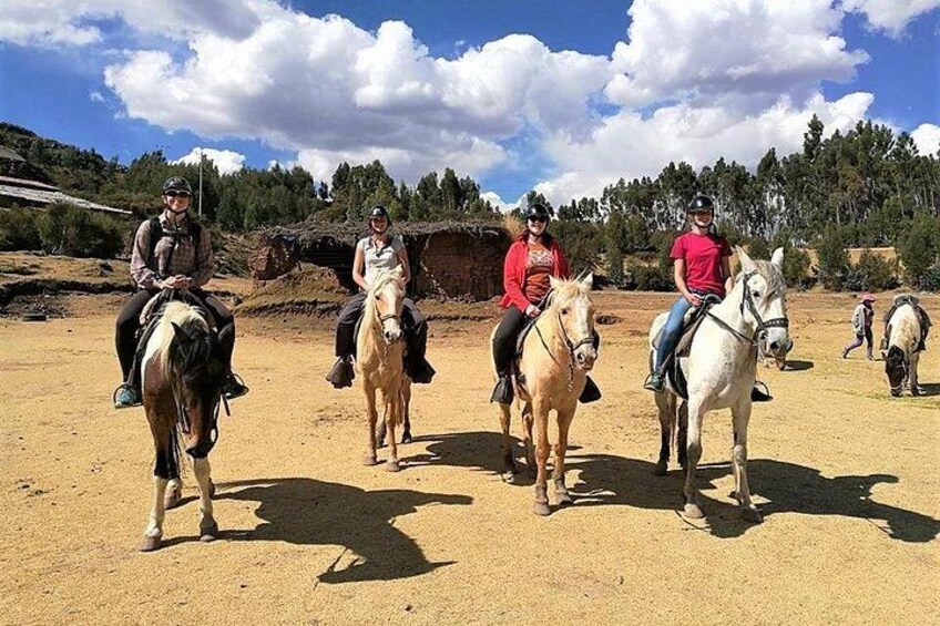 Four people riding horses on a dirt path with a thatched-roof structure in the background. 5