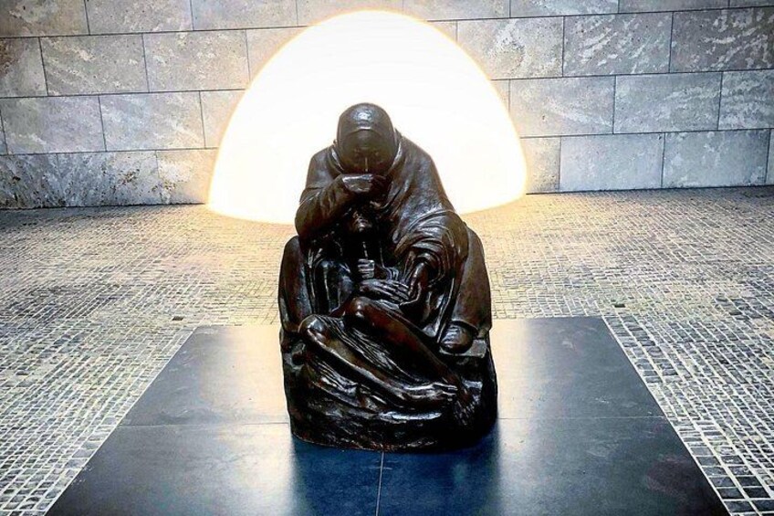 Una escultura de bronce muestra una figura sentada con un niño, frente a un suelo de baldosas y un gran aplique circular al fondo. 5