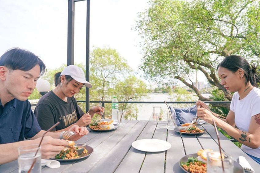 Three people dining outdoors at a wooden table with plates of food, a glass of water, and a bottle of water. 2