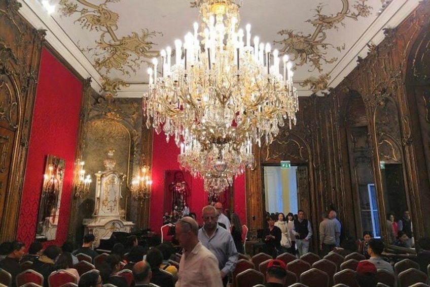 An ornate room with a large chandelier, red walls, and a crowd of people. 2
