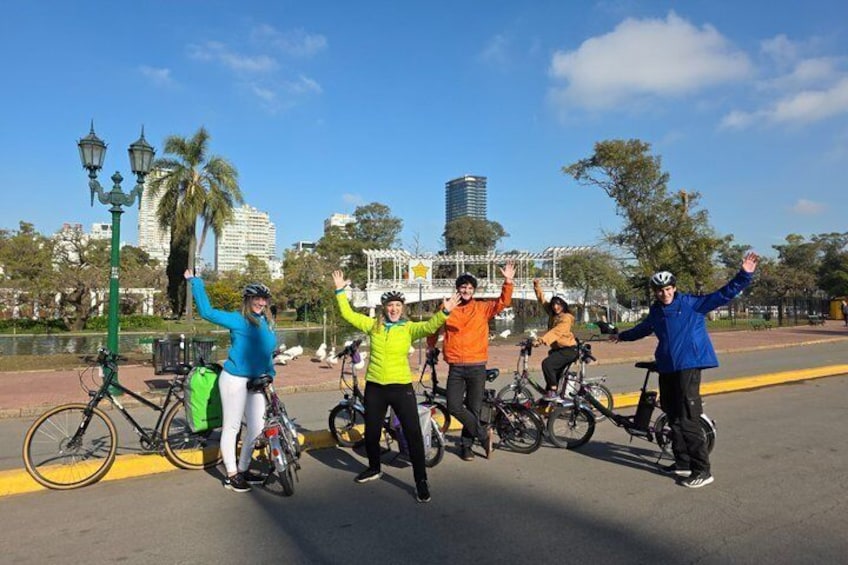 Un grupo de ciclistas en un parque con un lago, palmeras y edificios al fondo. 1