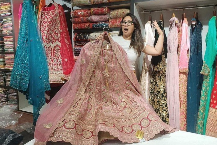 A person holding a pink embroidered fabric, with various colorful fabrics hanging in the background. 1