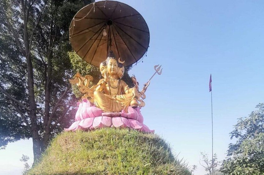 Een groot gouden standbeeld van een godheid met een drietand, zittend op een lotusvoetstuk, onder een grote parasol, omringd door groen. 2