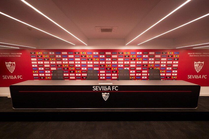 A press conference room with a long table, chairs, and a backdrop with logos. 3