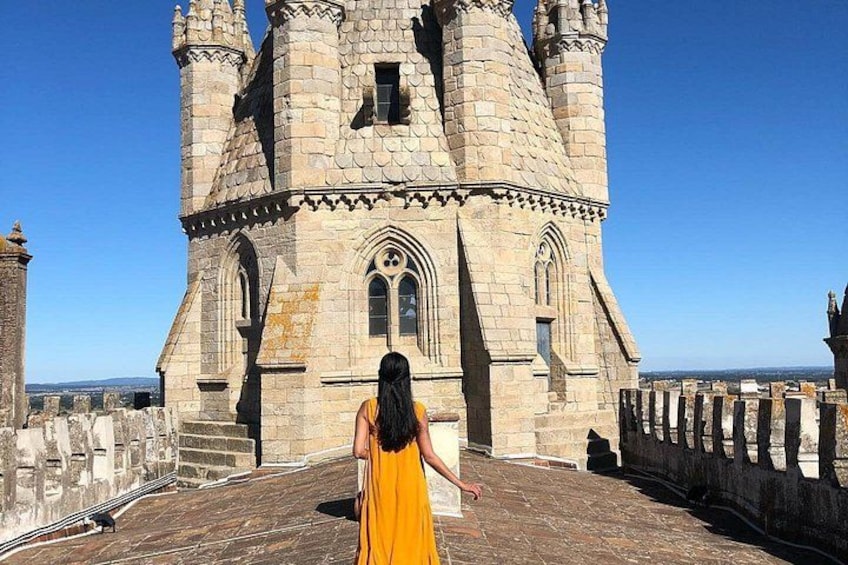 A historic stone building with multiple towers and arched windows, a person in a yellow dress, and a clear blue sky. 1