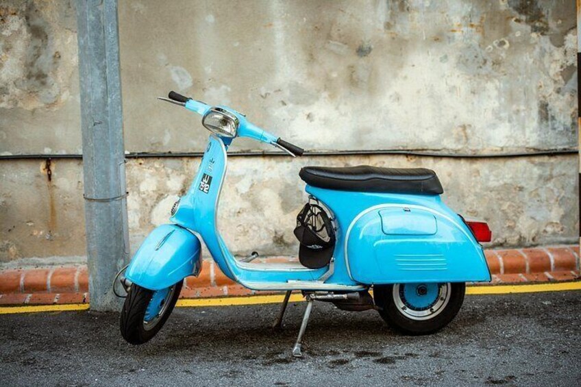 A blue vintage Vespa scooter parked on a street with a concrete wall and a metal pole in the background. 4
