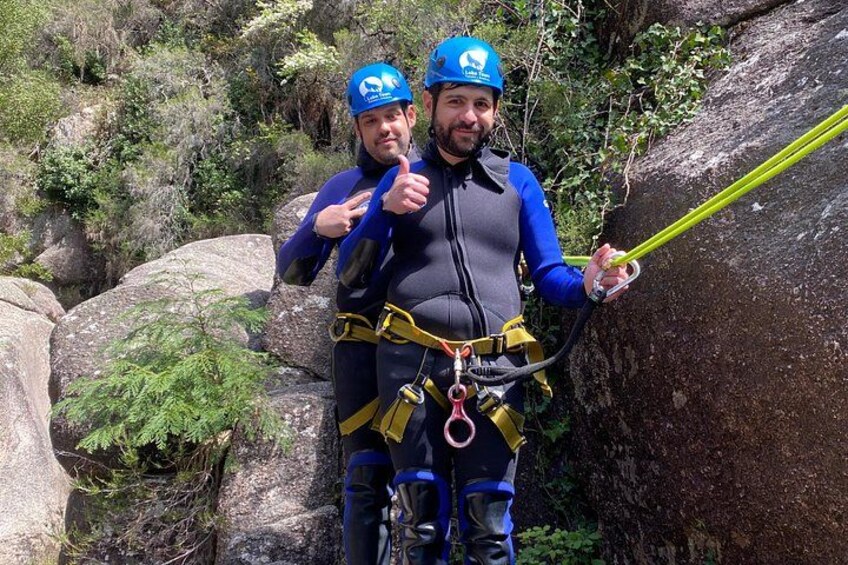 Deux personnes pratiquent la descente en rappel, équipées de harnais et de cordes, et sont entourées de gros rochers et de végétation luxuriante. 2