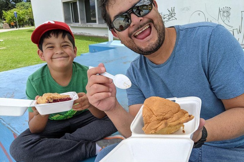 Two people sitting outdoors, each holding a food container with a sandwich. 3