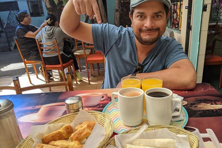 A person sitting at a table with a basket of croissants, two cups of coffee, and two glasses of orange juice. 1