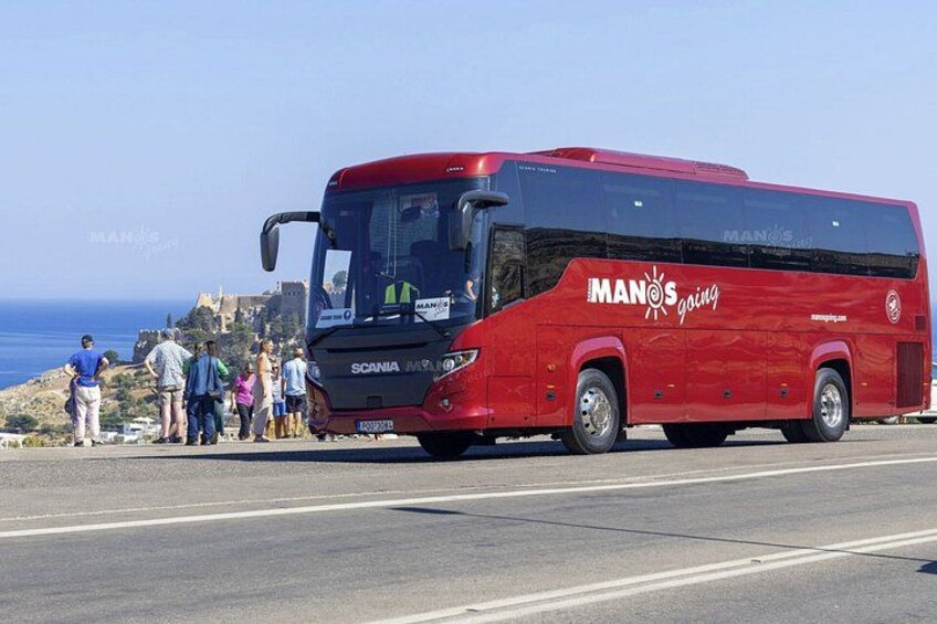 A red MANOS touring bus parked on a coastal road with people standing nearby. 2