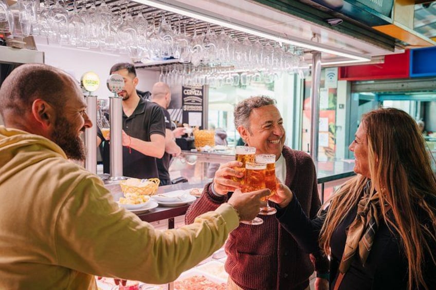 A bar setting with people toasting with beer glasses, a counter with food items, and hanging glasses above the bar. 4