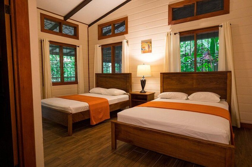Ein hölzernes Bett mit weiß-orange gestreifter Bettdecke, zwei Nachttische und ein Fenster mit Blick auf Grünfläche. 2
