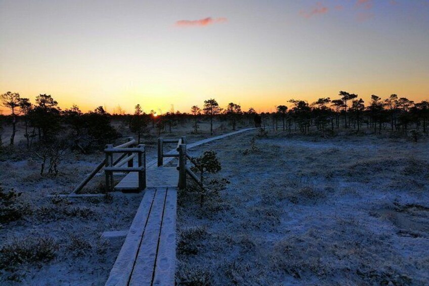 Una passerella in legno che si snoda attraverso un paesaggio innevato, con poca vegetazione e un tramonto sullo sfondo. 1