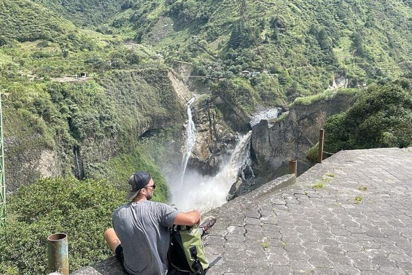 A person sitting by a waterfall with a backpack, surrounded by lush greenery and a rocky terrain. 2