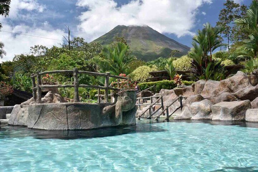A pool with a wooden bridge and rocks, surrounded by lush greenery and a mountain in the background. 1