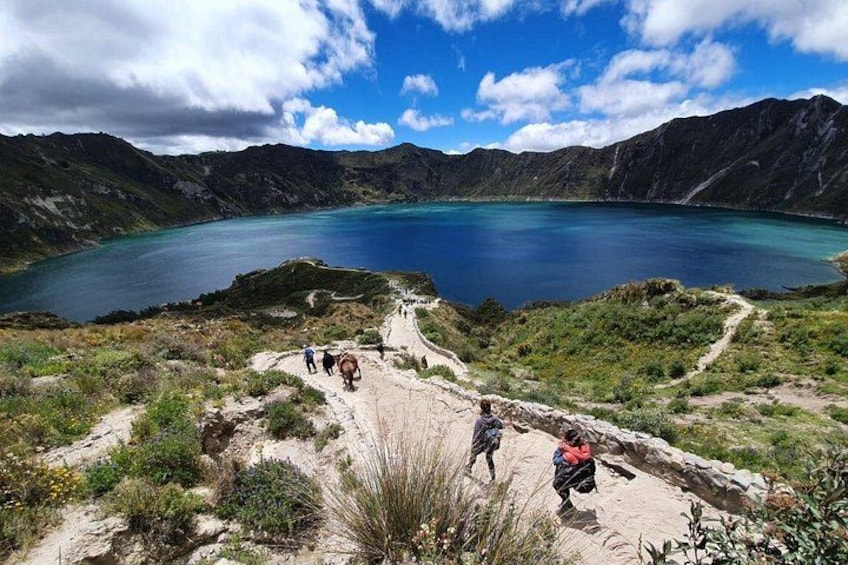 A mountainous landscape with a clear blue lake, a dirt path, and hikers. 1