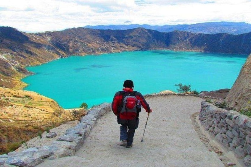 A hiker with a red backpack walking on a dirt path towards a turquoise lake surrounded by mountains. 3