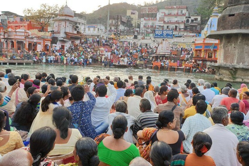 A crowd of people gathered by a river, with buildings and a temple in the background. 2