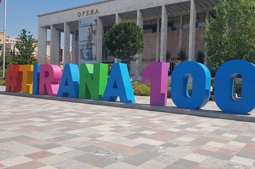 A colorful sign with the text '#TRANA100' in front of a building labeled 'OPERA.' 4