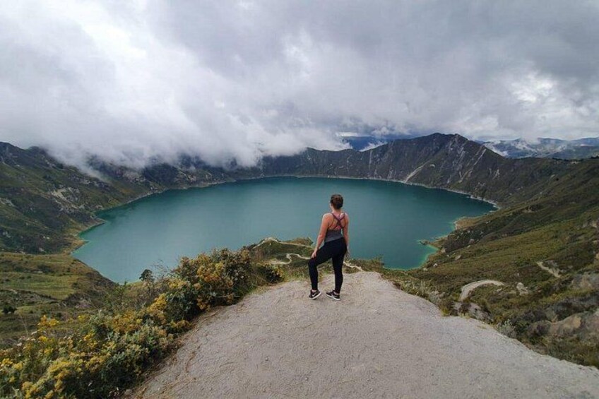 A person standing on a mountain path overlooking a turquoise lake surrounded by mountains and clouds. 5