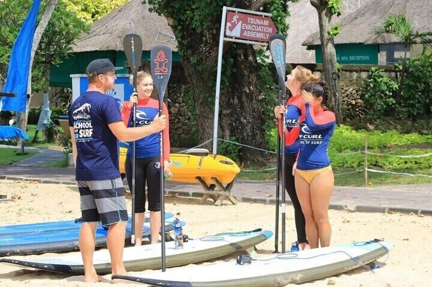People on a beach with paddleboards, a sign for a tsunami evacuation assembly, and a yellow boat in the background. 3