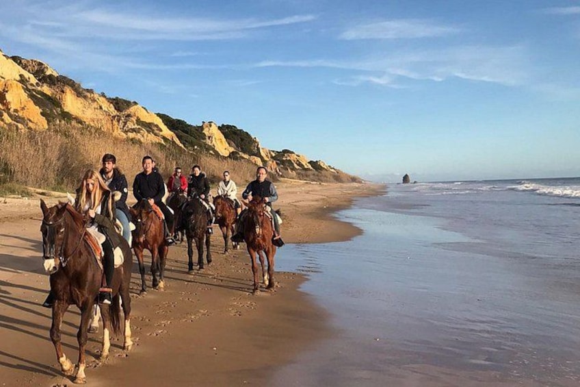 A group of people riding horses along a beach with cliffs in the background. 1