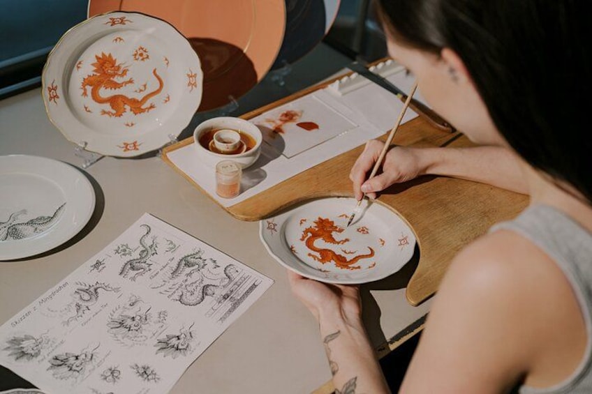 A person painting on a plate with a dragon design, surrounded by other plates and a sketchbook with dragon drawings. 2