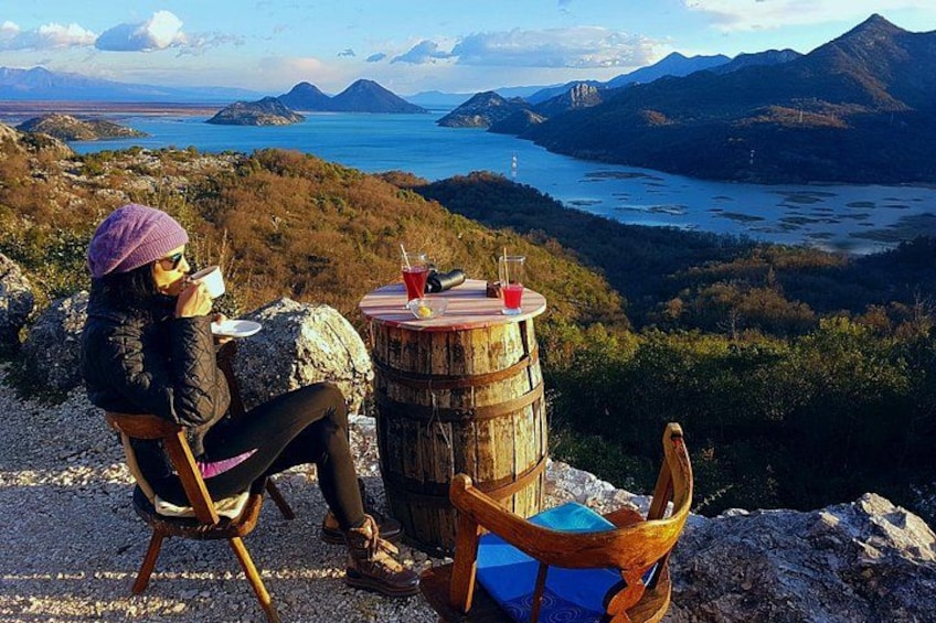 A person sitting on a chair, holding a cup, with a scenic view of mountains and a lake in the background. 2