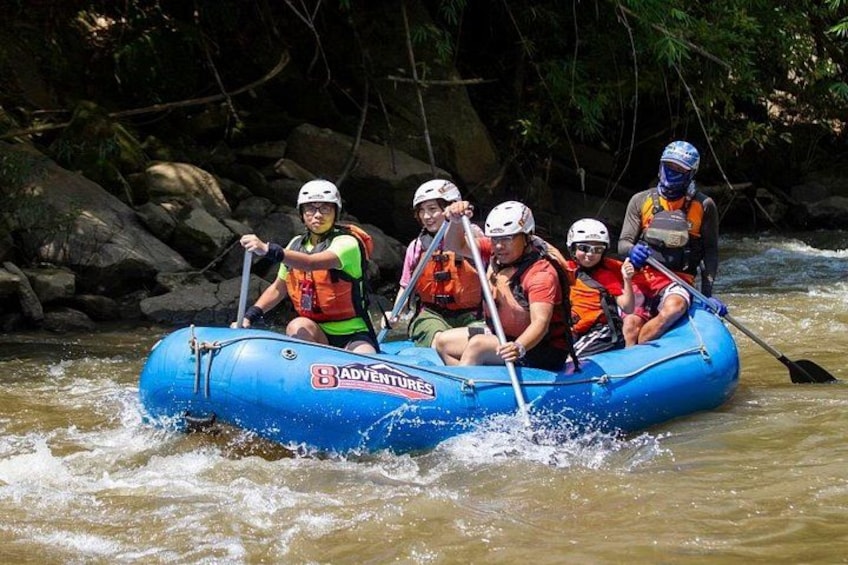 Een groep mensen die op een opblaasbare, blauwe raft stroomafwaarts vaart, de raft is beletterd met 'ADVENTURES', en navigeert door een rivier omringd door rotsen en bomen. 2