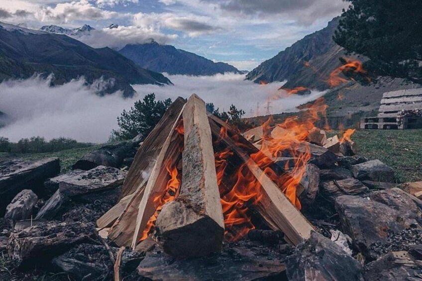A campfire with logs and flames, surrounded by rocks and a mountainous landscape with clouds. 3
