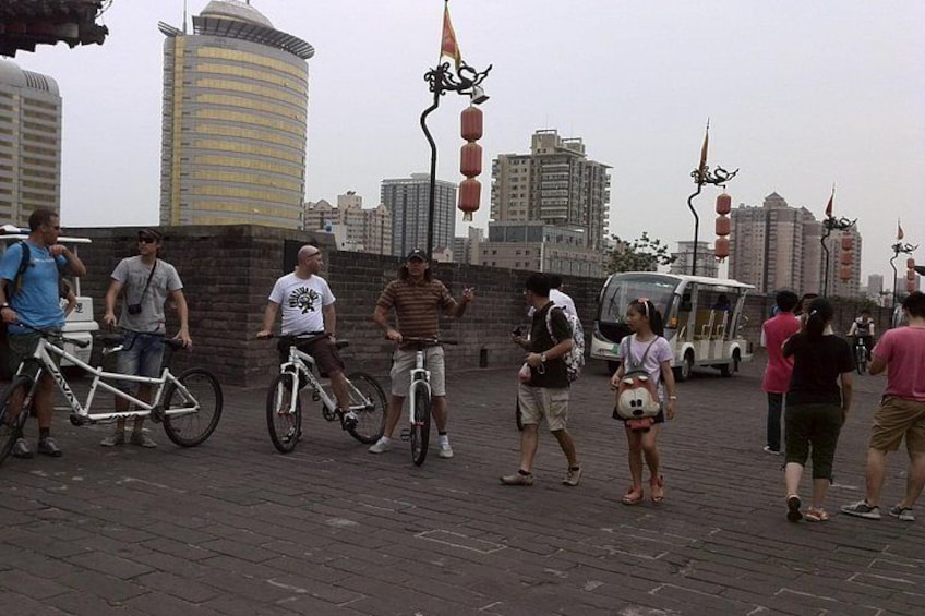 People standing and walking on a paved area with bicycles, a bus, and buildings in the background. 4