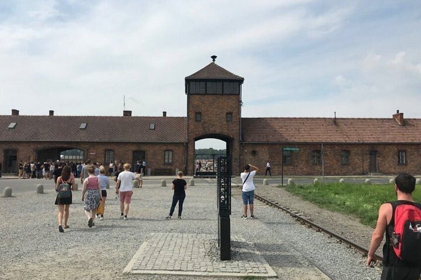 Ein historisches Gebäude mit einem Wehrturm, einer gepflasterten Fläche mit Passanten und einer Bahngleis. 2