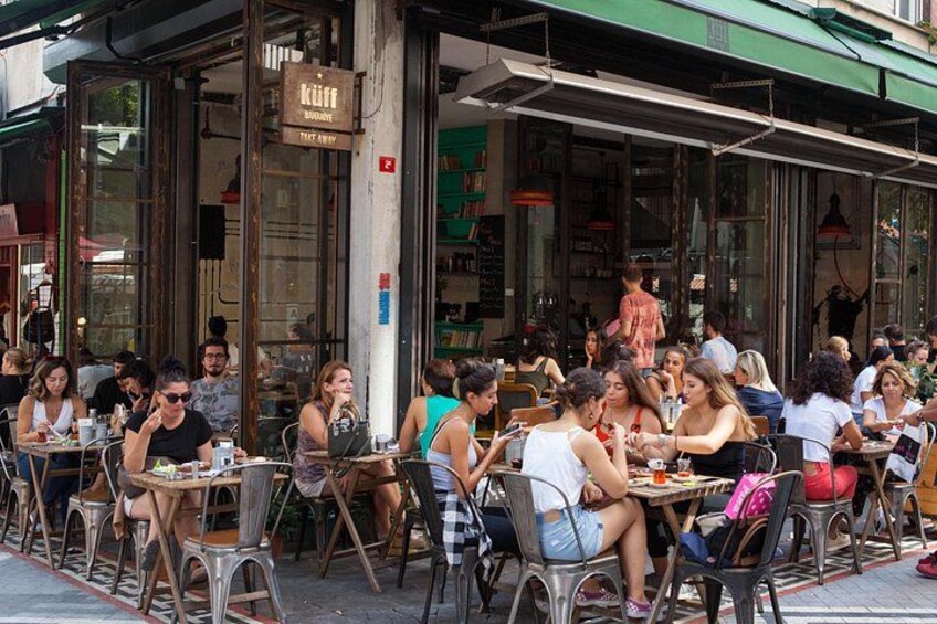 Ein belebtes Straßencafé mit Gästen an Tischen, einer grünen Markise und einem Schild über dem Eingang. 3