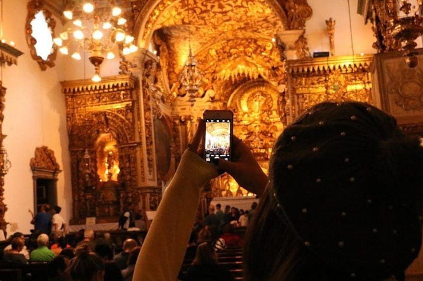 A person taking a photo inside a richly decorated church with ornate golden details and a large chandelier. 4